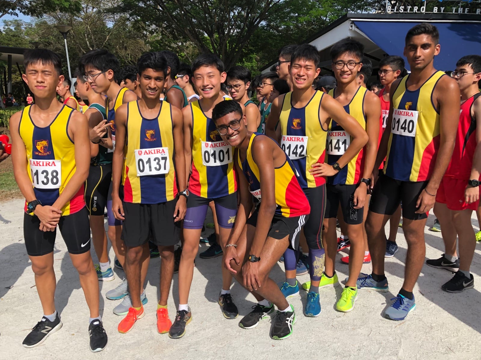 Six teenage male runners in yellow and blue Akira race bibs pose together outdoors before a cross-country race.
